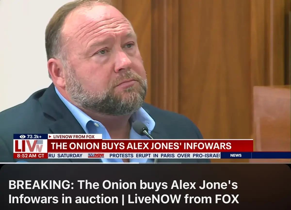 A man with a beard and receding hairline, wearing a suit and blue shirt, looks off to the side while sitting at a desk with…