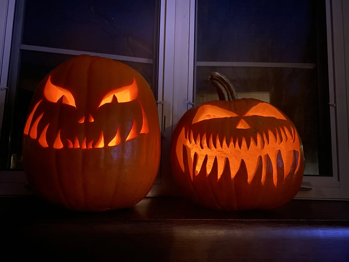 Two carved Halloween pumpkins sit side-by-side on a windowsill at night, glowing from within. The pumpkin on the left has a…