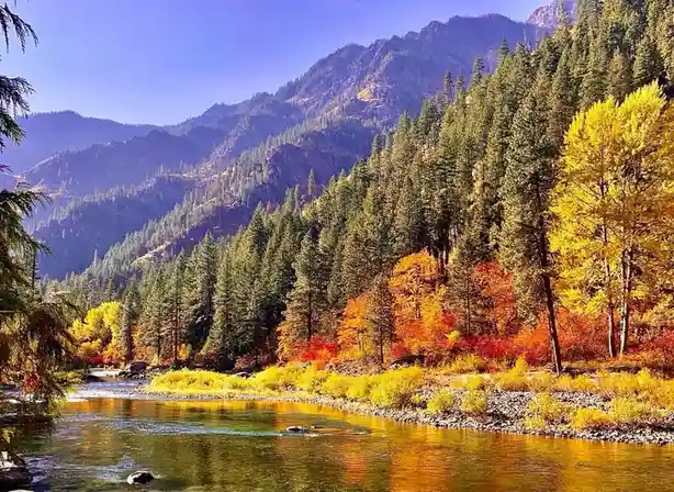 A vibrant autumn landscape featuring a river flowing through a valley with mountains in the background. Trees display bright…