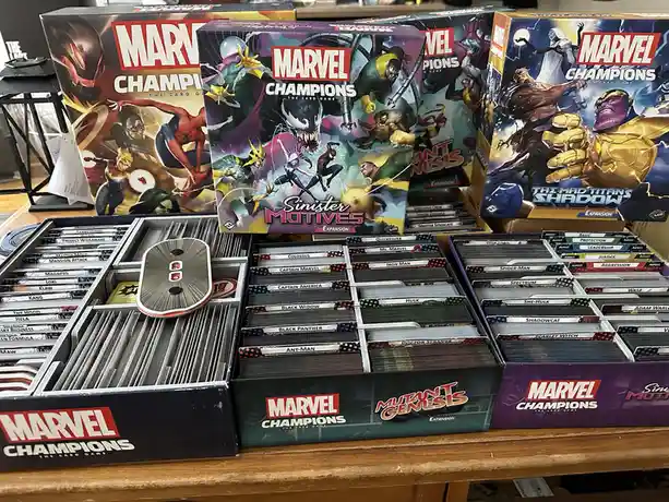 A collection of Marvel Champions: The Card Game boxes and card storage trays are displayed on a wooden shelf. The boxes…