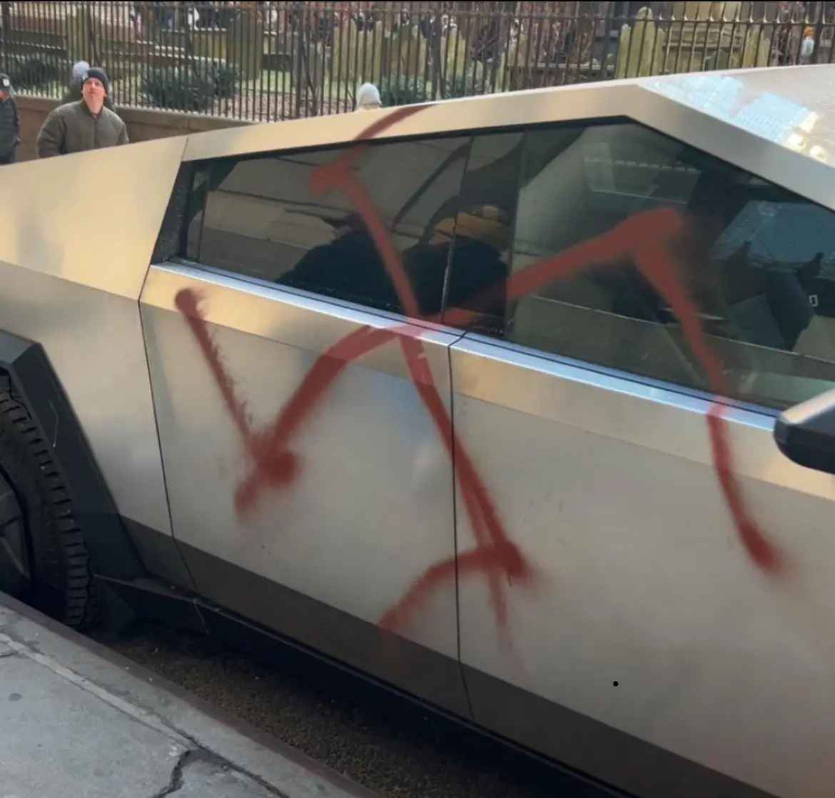a silver, angular electric vehicle, resembling a Tesla Cybertruck, has a large red spray-painted 'X' across its…