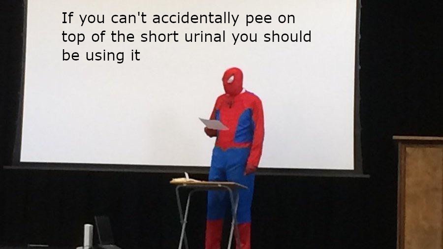 A person in a full Spider-Man costume stands at a lectern in front of a large projection screen, holding a paper. The screen…