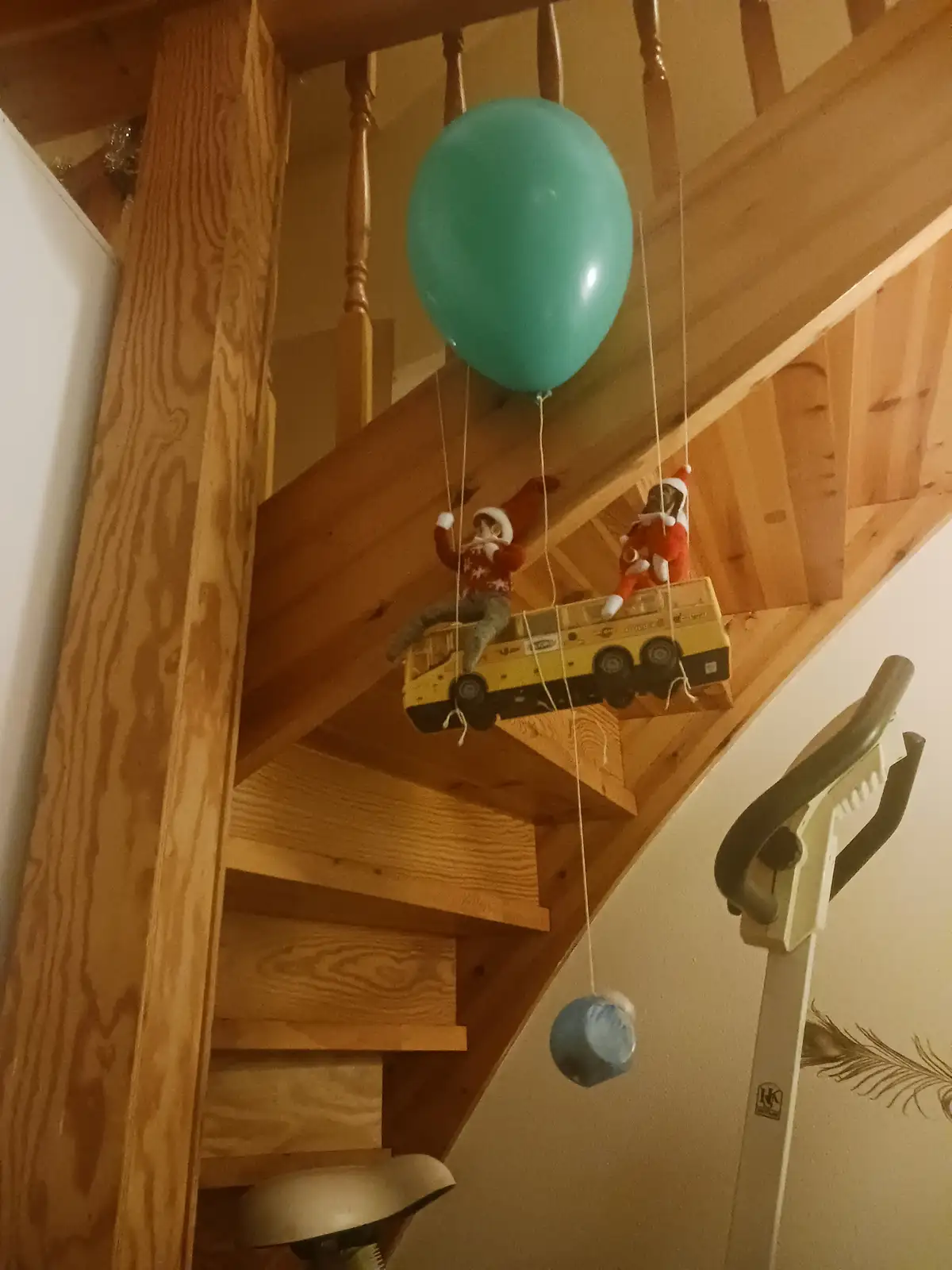 A teal balloon is tied to a toy yellow bus with two elf figurines wearing red and white outfits, suspended by strings from a…