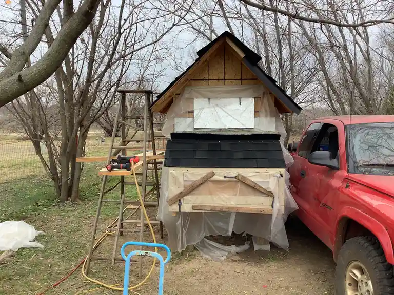 An unfinished wooden doghouse with a black shingled roof and plastic sheeting covering parts, sits next to a red pickup…