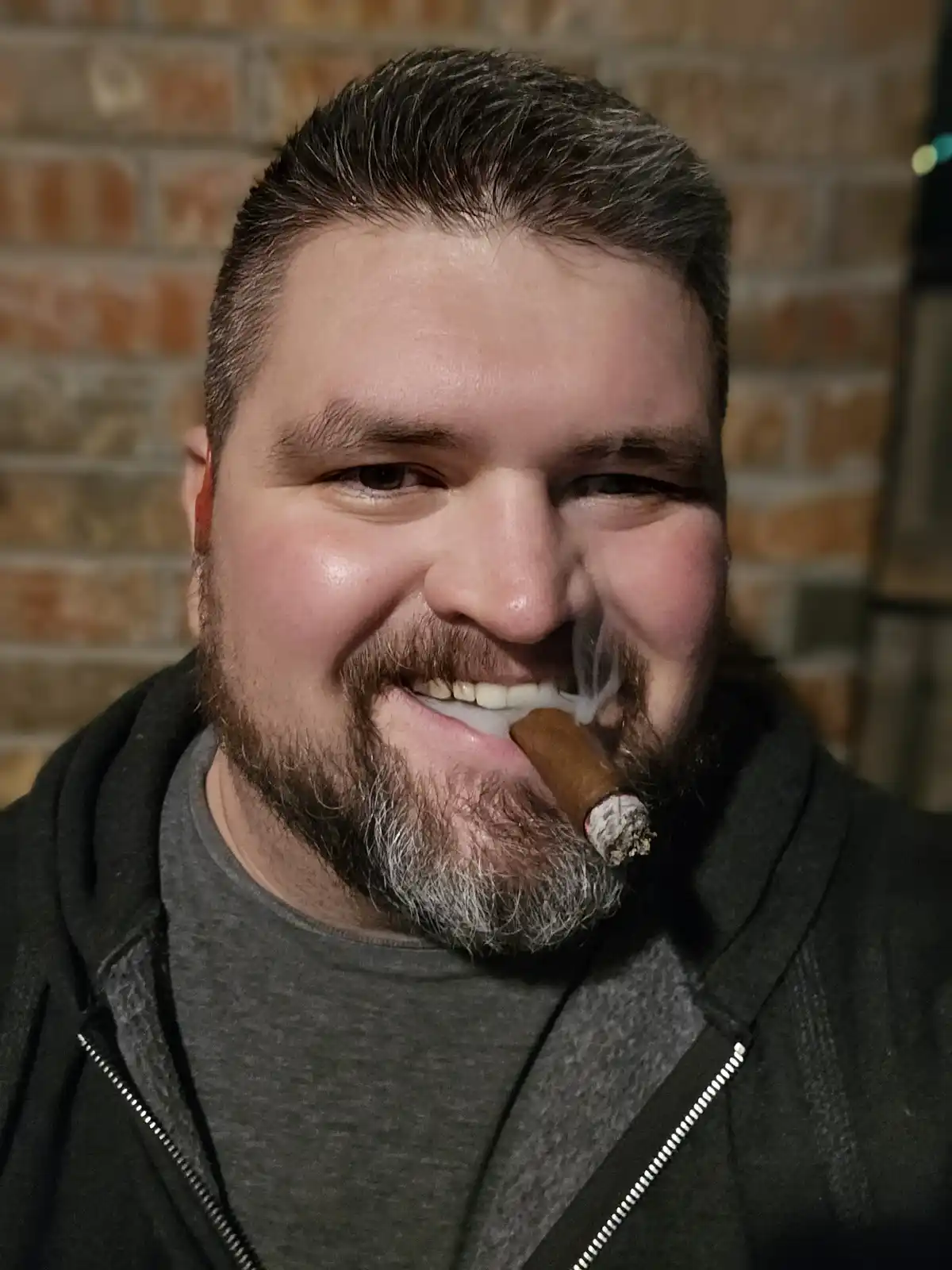 a man with a beard and short hair smiles while holding a lit cigar in his mouth, smoke visible, against a brick wall…