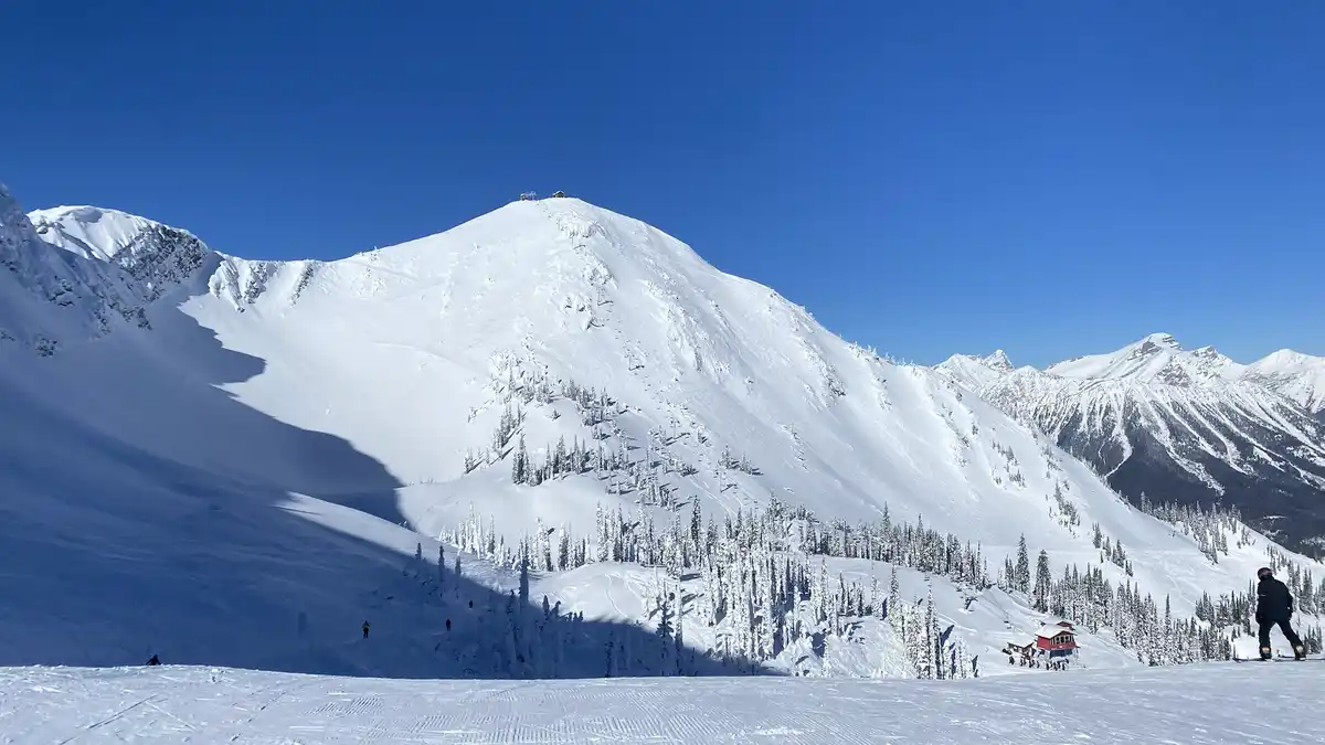 A wide-angle photo of a snow-covered mountain range under a clear blue sky, with a skier in the foreground on a groomed…