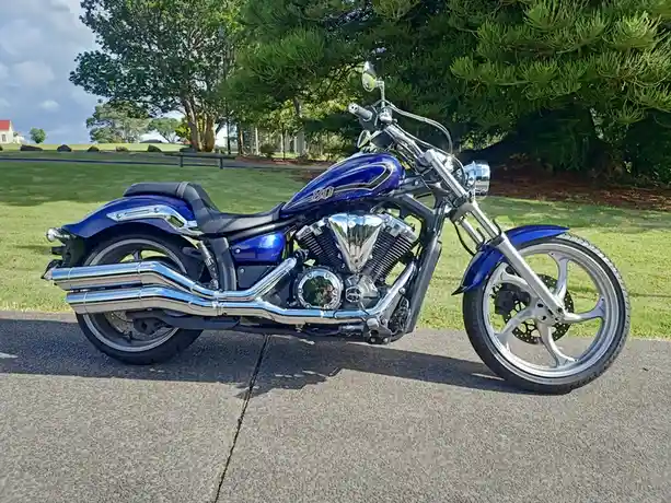 A shiny blue Yamaha Stryker motorcycle is parked on a paved path in a grassy park with trees in the background. The bike…