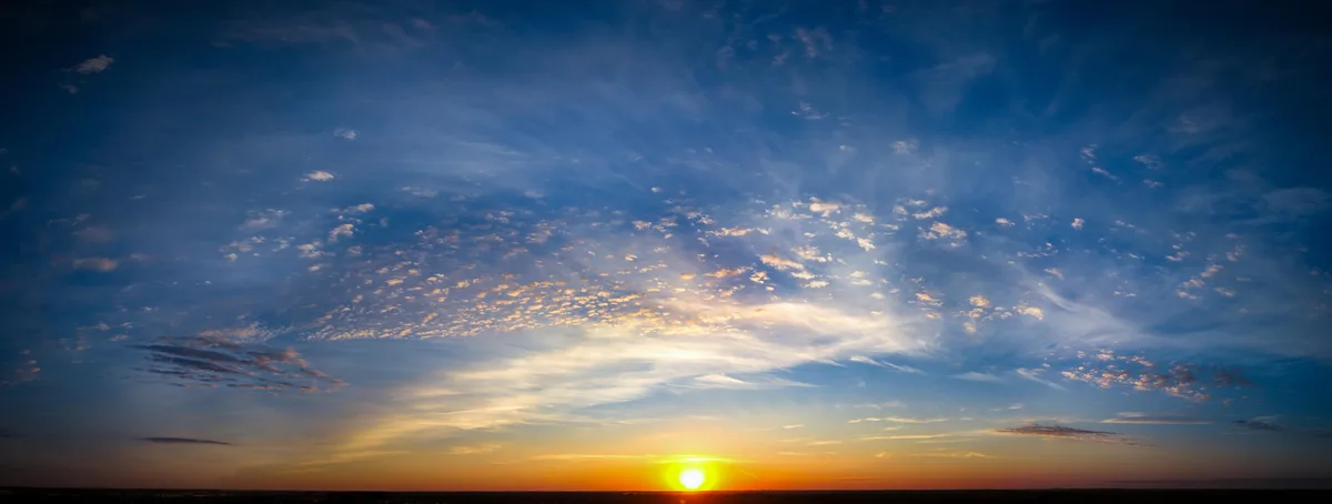 A panoramic photograph captures a vibrant sunset with the sun low on the horizon, casting orange and yellow hues across the…