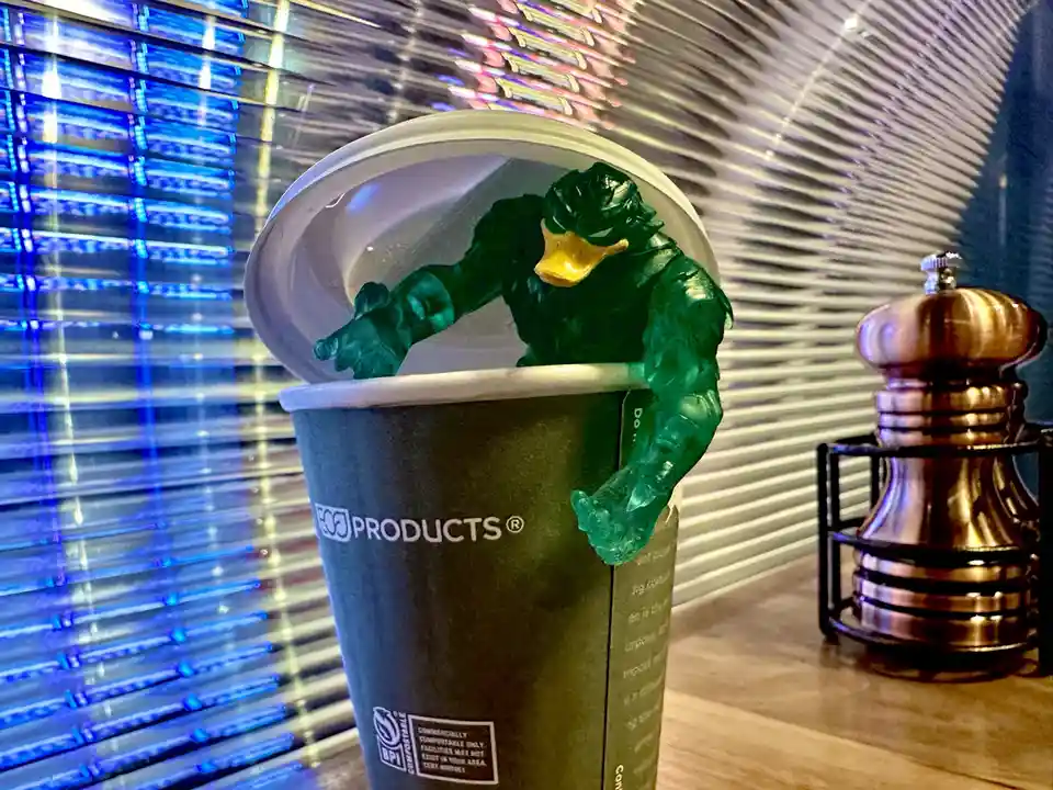 A green, muscular duck action figure is positioned as if emerging from a dark green disposable coffee cup with a white lid…