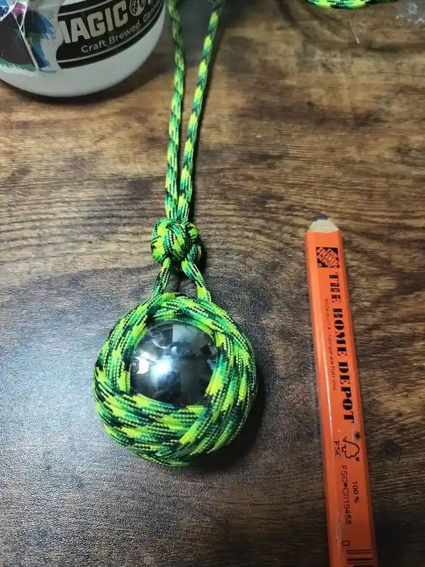 A handmade paracord pendant with a circular mirror embedded in its center rests on a wooden surface next to an orange Home…