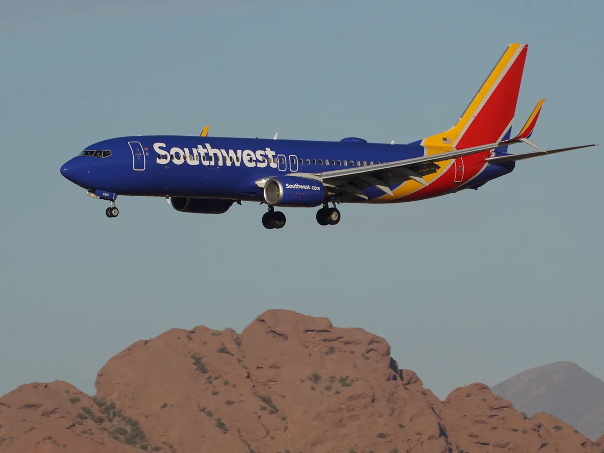 A blue Southwest Airlines Boeing 737 with red and yellow tail is flying low over reddish-brown desert mountains with its…