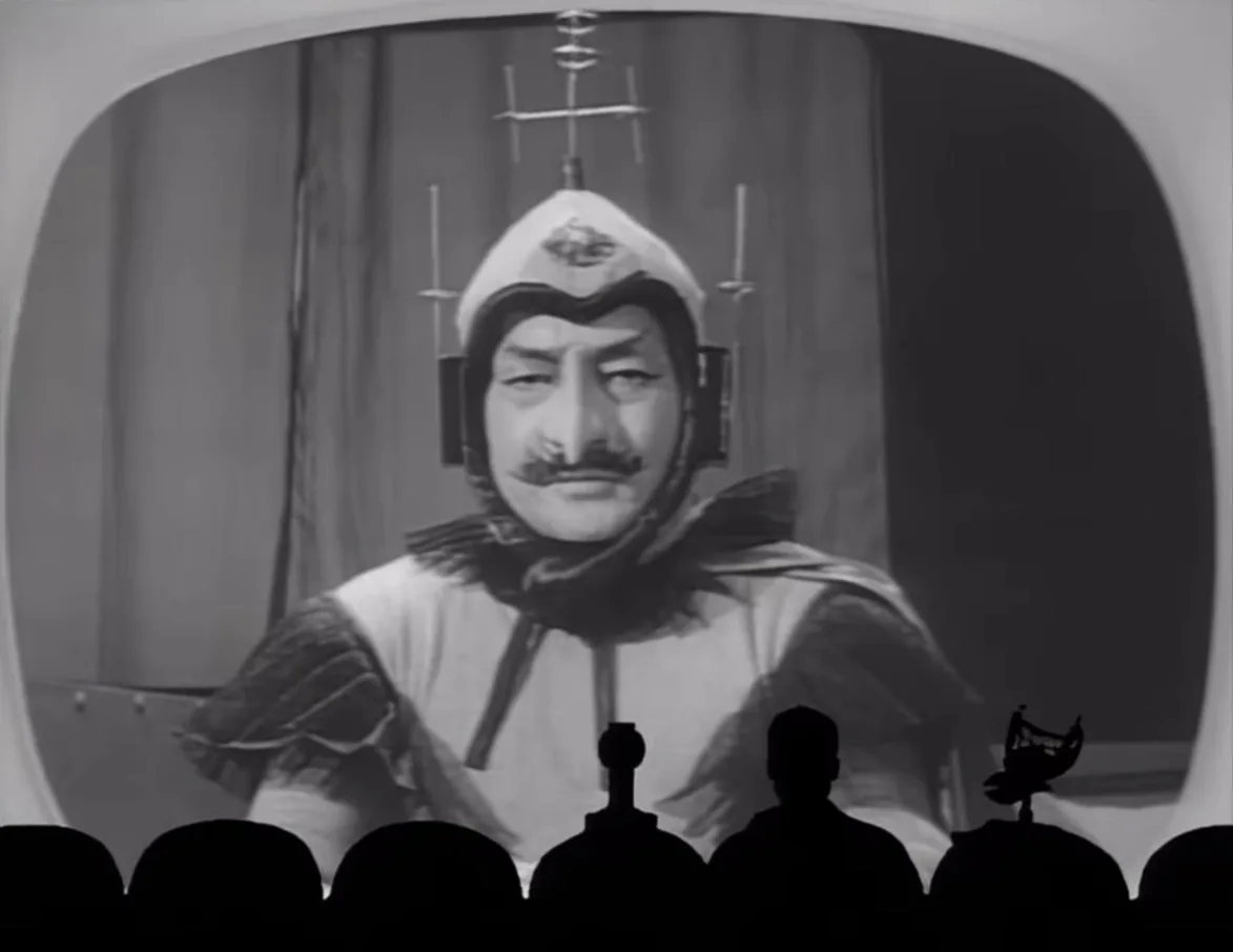 A black-and-white image shows a man in a futuristic helmet with antennae, viewed on a screen from the perspective of an…