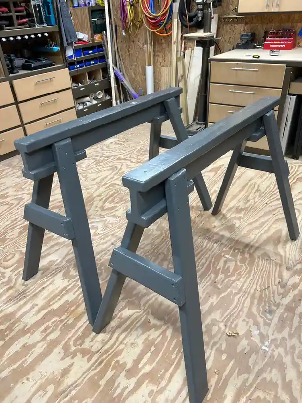 Two identical gray-painted wooden sawhorses stand side by side on a plywood floor in a cluttered workshop, surrounded by…