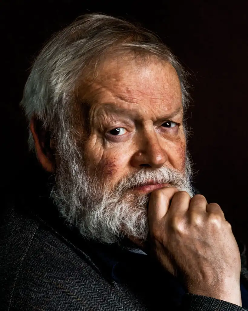 a close-up portrait of an older man with a white beard and gray hair, wearing a dark herringbone jacket, looking intently at…