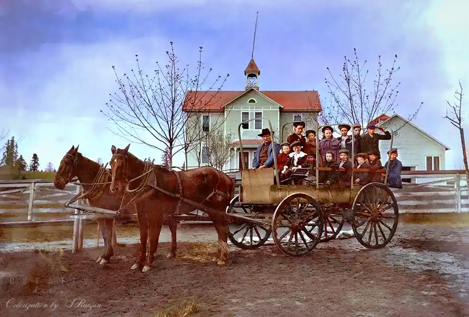 An old, colorized photograph shows a large group of children and adults sitting in a horse-drawn wagon pulled by two brown…