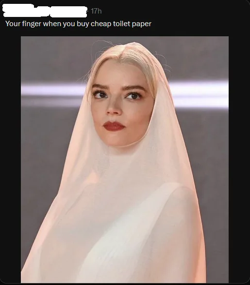 A woman with platinum blonde hair and dark lipstick wears a sheer white veil draped over her head and shoulders, looking…