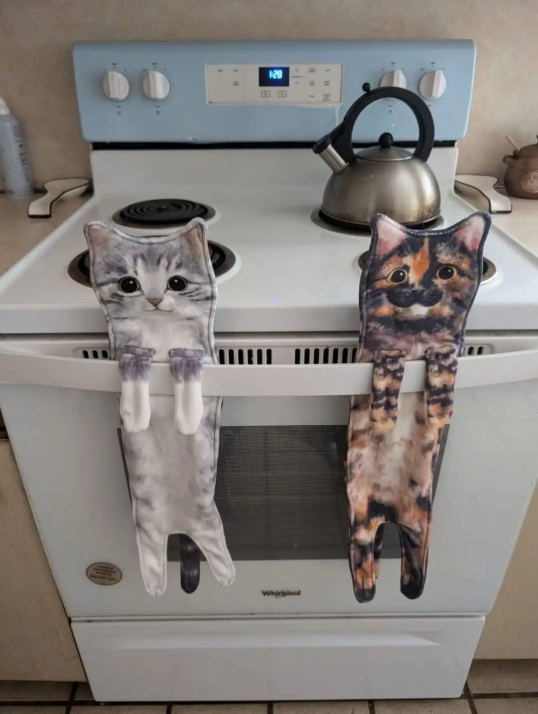 Two cat-shaped oven mitts hang from the handle of a white Whirlpool stove, one gray tabby and one calico, looking directly…