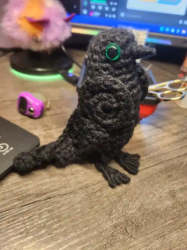 A handmade black crocheted bird with one large green eye sits on a wooden desk, surrounded by crafting tools and…