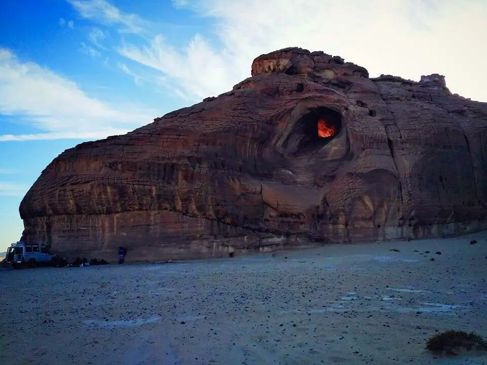 a large desert rock formation with a natural hole resembling an eye glowing with orange fire, with a white vehicle and…