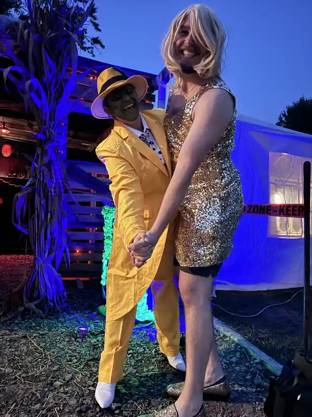 Two people pose together at an outdoor event under blue lighting; one wears a full green-face mask, yellow suit, and fedora…