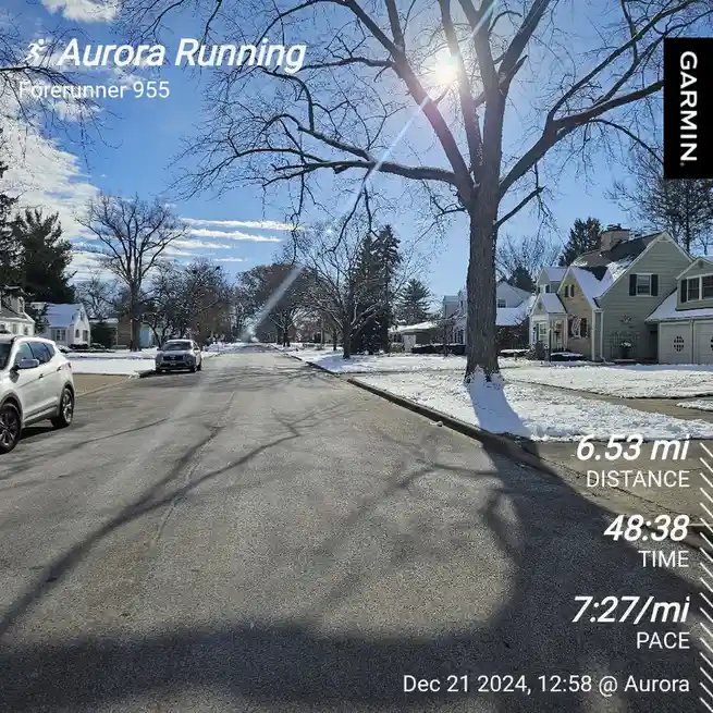 A screenshot from a Garmin Forerunner 955 running watch shows a winter street scene with snow on lawns and bare trees under…