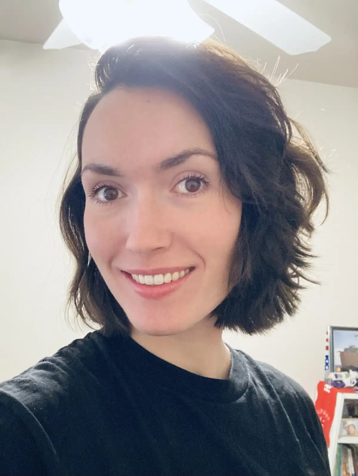 A selfie of a woman with short, dark, wavy hair smiling broadly at the camera. She wears a black t-shirt and is indoors…