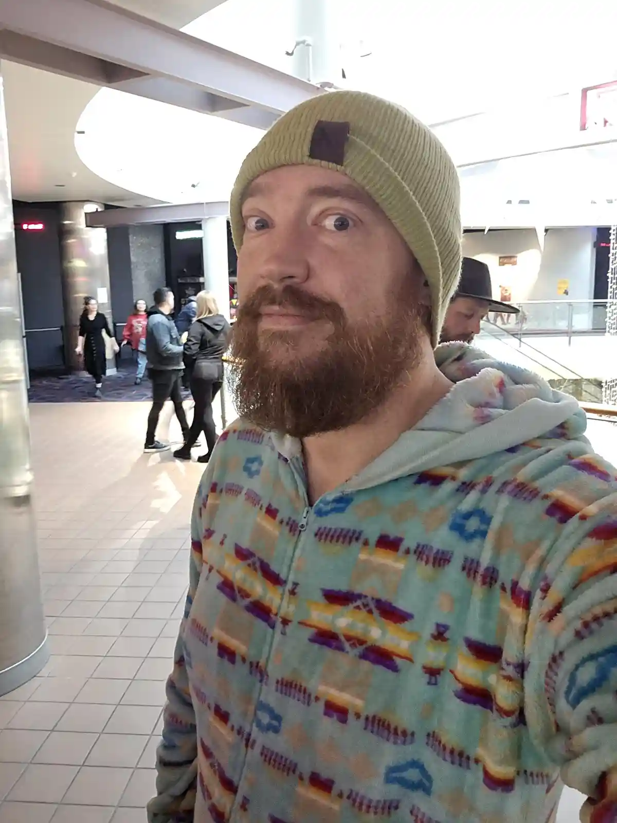 A man with a reddish beard and a light green beanie stares directly at the camera with wide, startled eyes while wearing a…