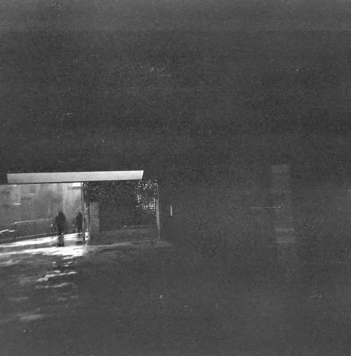 A grainy, black-and-white photo shows two silhouetted figures walking under a lit awning at night on a wet street, with the…