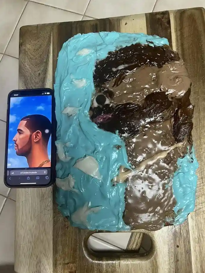 A cake decorated with blue and brown frosting to resemble a portrait of Drake, placed next to a smartphone displaying…