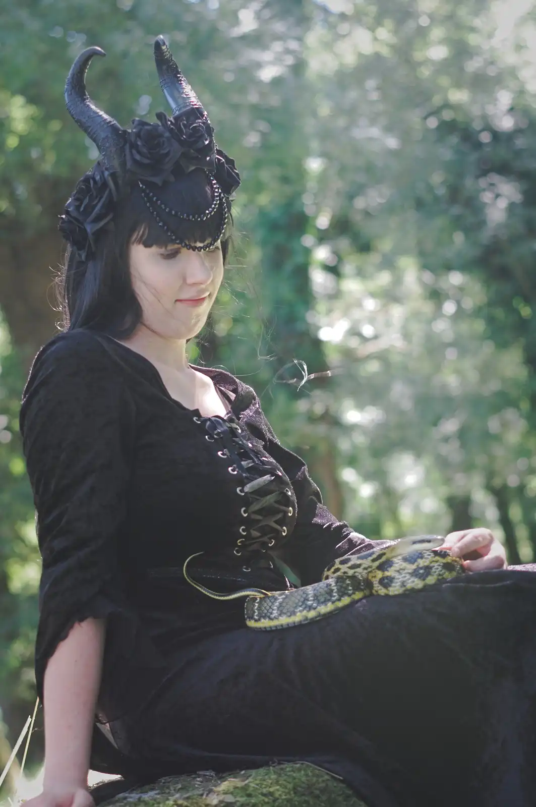 A woman dressed in black gothic attire with ornate horns and black roses on her head sits outdoors in a forest, calmly…