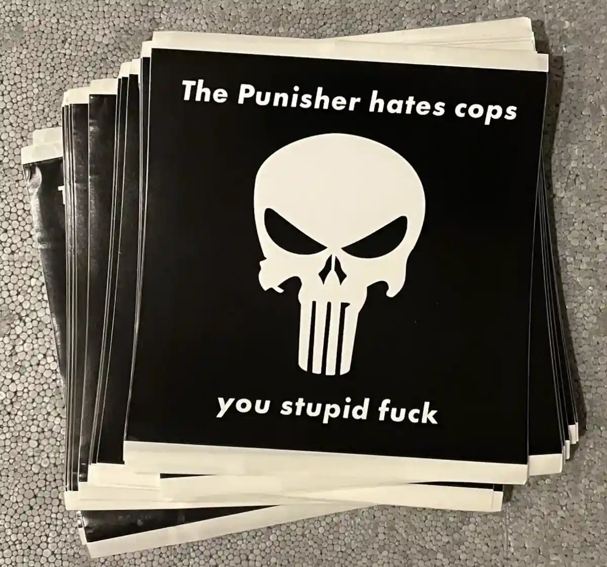 A stack of black square stickers with white borders, each featuring the iconic white skull logo of The Punisher. Above the…