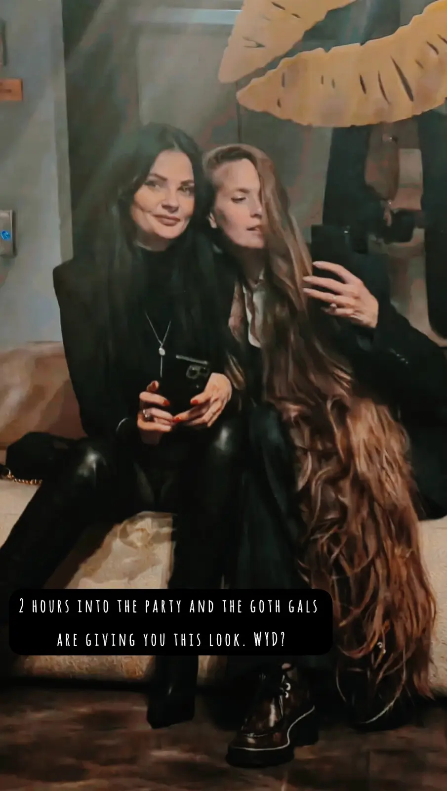 Two women dressed in dark, gothic-style clothing sit on a couch, holding phones, with one looking at the camera with a bored…