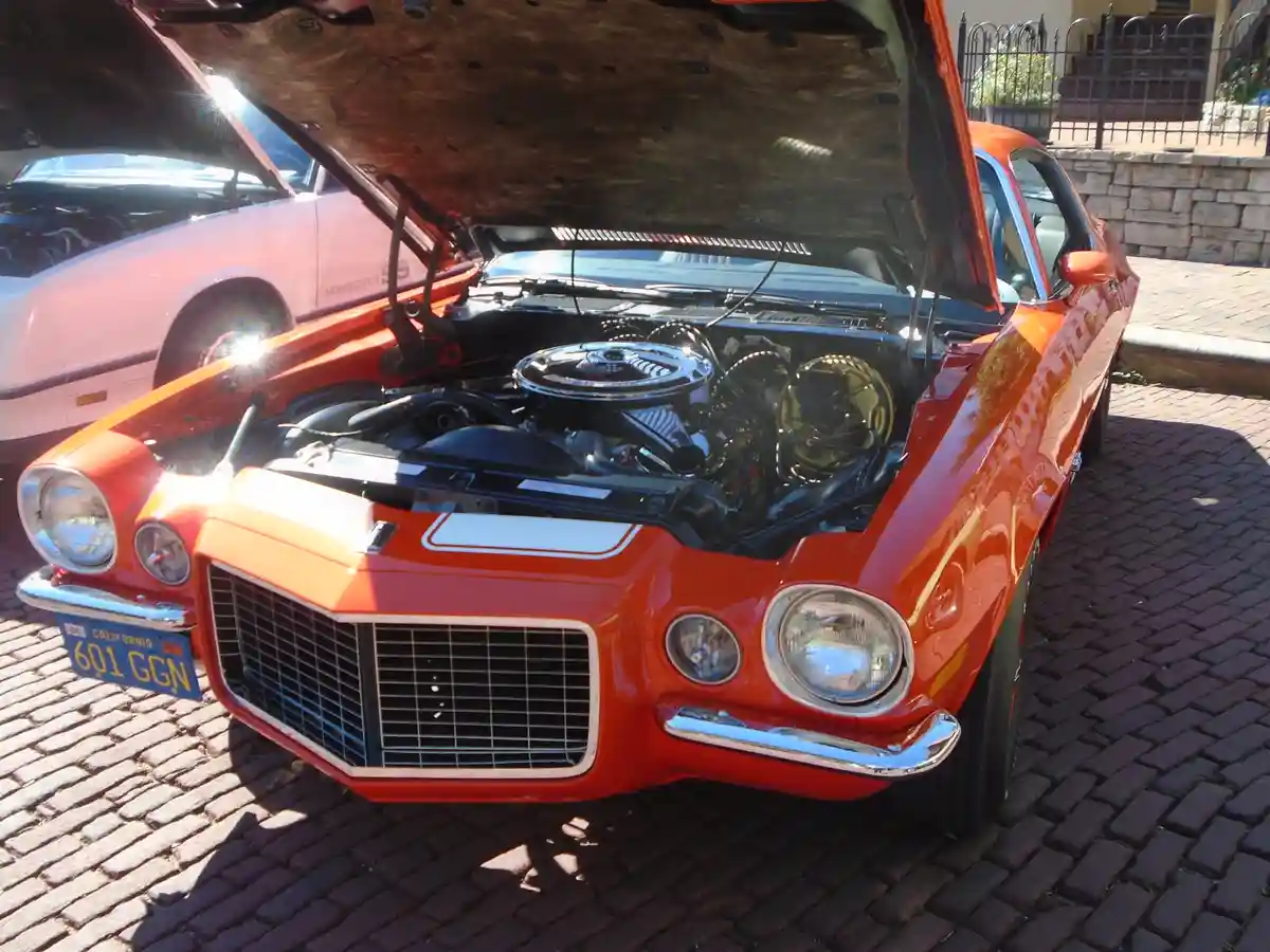 A bright orange classic muscle car with its hood open, revealing a detailed engine, parked on a brick driveway. The car has…