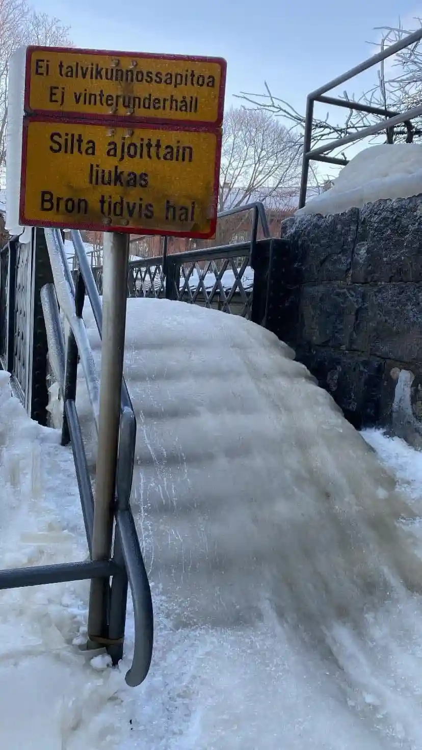 A yellow warning sign with red borders stands beside a steep, icy bridge ramp covered in snow and ice. The sign, in Finnish…