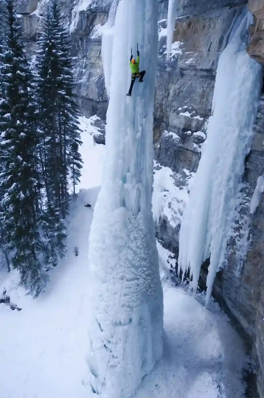 An ice climber in a bright green jacket and orange helmet ascends a massive, sheer frozen waterfall in a snowy mountain…