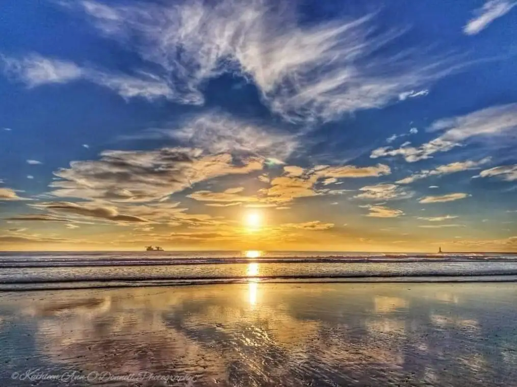 A vibrant, high-contrast photograph of a beach at sunrise or sunset, with the sun low on the horizon casting golden light…
