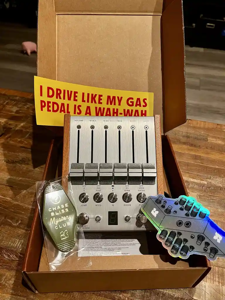 An open cardboard box displays a Chase Bliss audio effects pedal with multiple knobs and sliders, accompanied by a green…