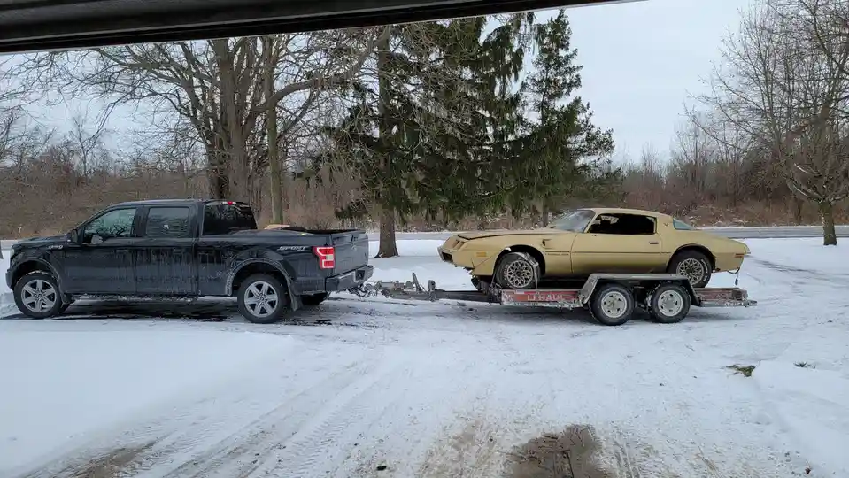 A black modern Ford F-150 pickup truck is towing a trailer with a gold 1970s Pontiac Trans Am muscle car in a snowy outdoor…