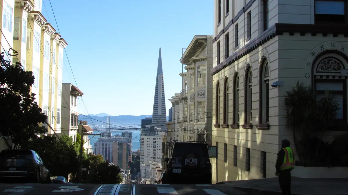 A photo of a steep San Francisco street with Victorian buildings, the Transamerica Pyramid in the distance, and a person in…