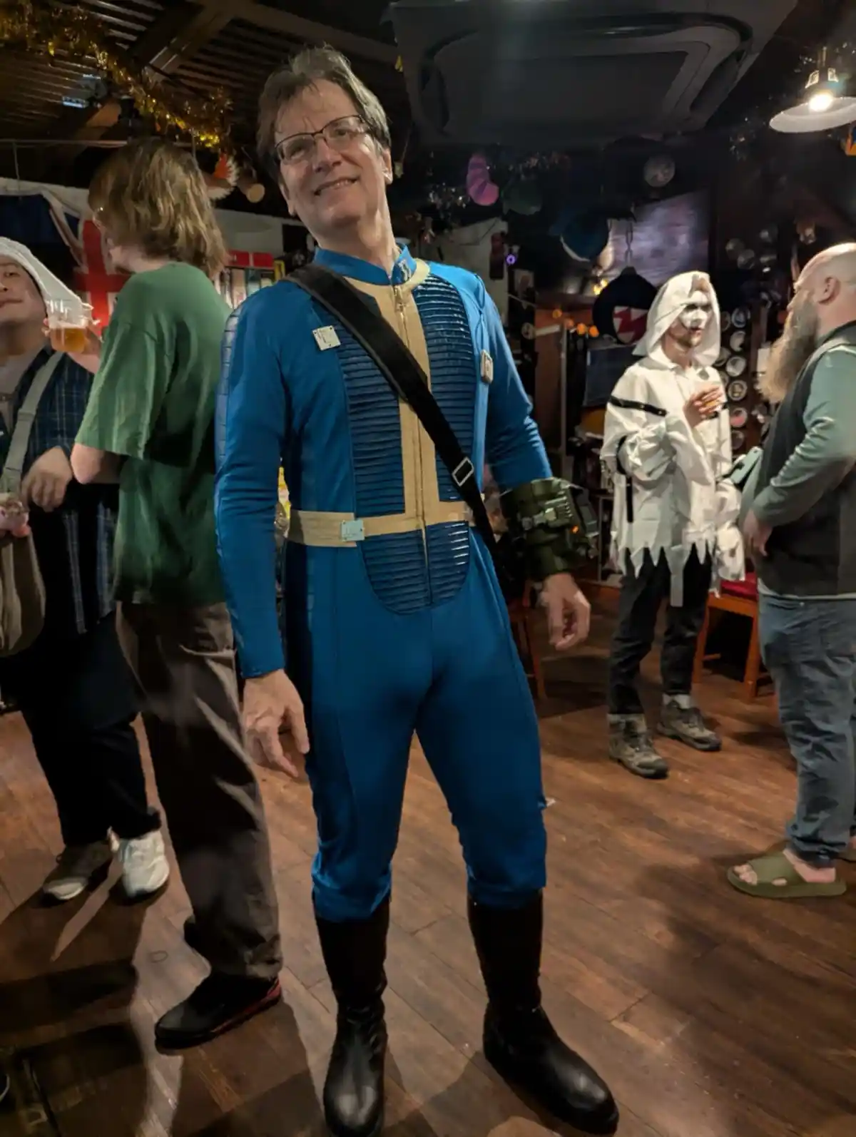 A man in a blue and tan Fallout power armor costume smiles at the camera while standing in a crowded room with other people…