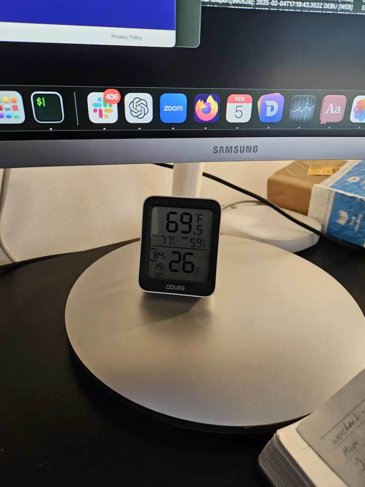 A Govee digital thermometer and hygrometer displays 69.5°F and 26% humidity on a desk in front of a Samsung monitor showing…