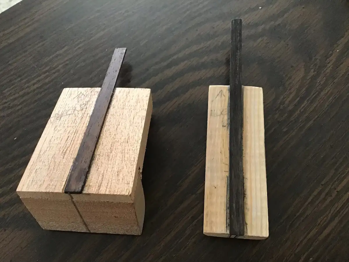 Two wooden blocks sit on a dark wood surface, each with a dark, narrow strip embedded vertically. The block on the left has…