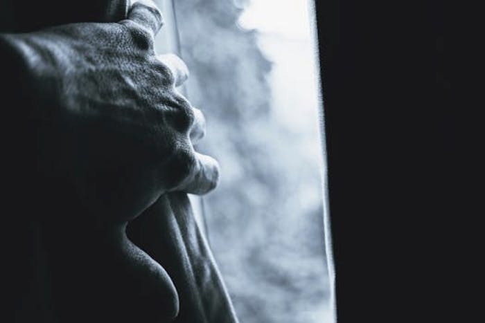 A close-up, high-contrast black-and-white photo shows a hand gripping the edge of a window frame, with bright light and…