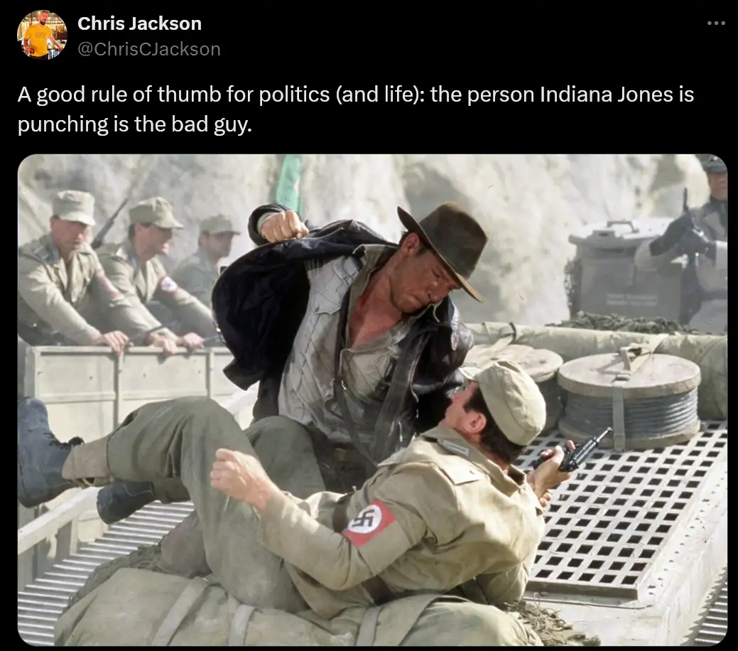 Indiana Jones, wearing a brown fedora and leather jacket, is punching a soldier in a tan uniform with a swastika armband…
