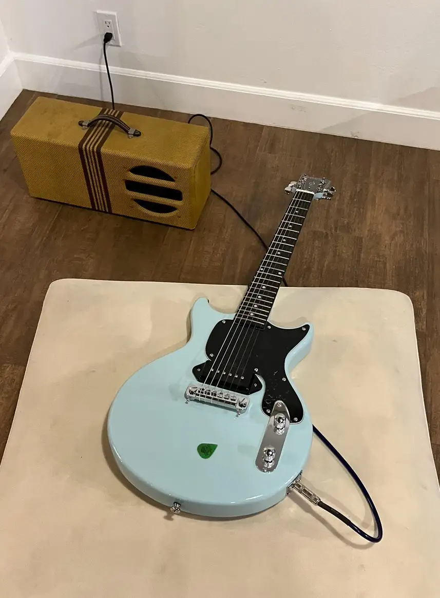 A light blue electric guitar with a black pickguard and silver hardware rests on a beige mat on a wooden floor. In the…