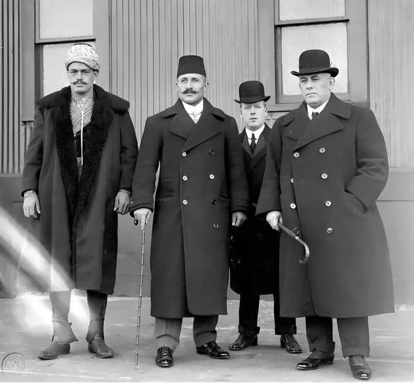 A black-and-white historical photo shows four men in long coats standing outdoors. From left to right: a man in a patterned…