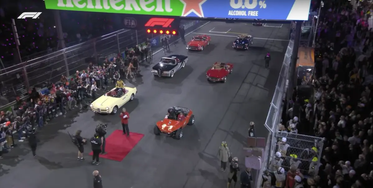 An overhead view of a Formula 1 track at night during a classic car parade, with several vintage cars driving past crowds…
