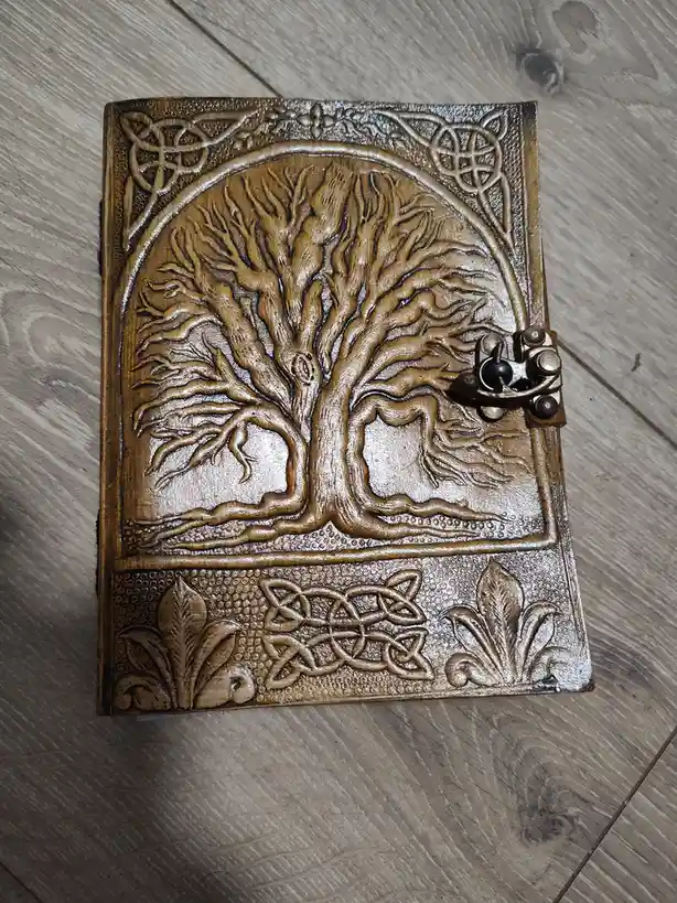 A top-down photo of a brown leather-bound journal with intricate embossed designs, featuring a large tree of life with roots…