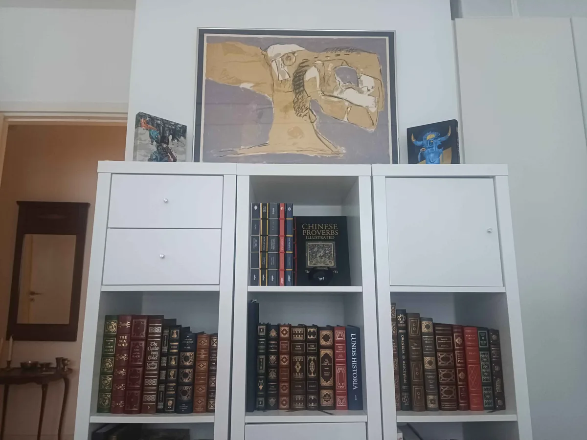A white bookshelf filled with numerous classic and decorative hardcover books, with a large abstract painting and two small…