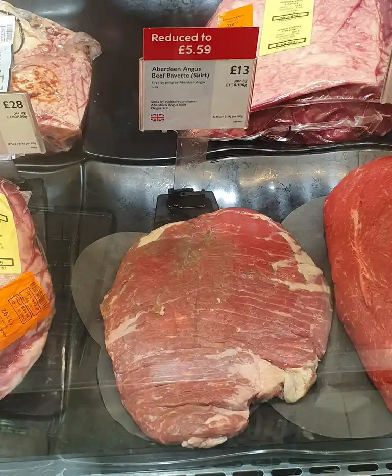A close-up photo of a large, raw Aberdeen Angus beef bavette (skirt) steak displayed in a supermarket meat case, with a red…